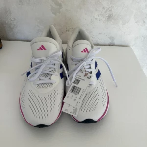 Vita Adidas Supernova 2 löparskor - Säljer ett par vita Adidas Supernova 2 löparskor med blå och rosa detaljer. Skorna har snörning och en sula med svart och blått mönster. Perfekta för löpning och träning.