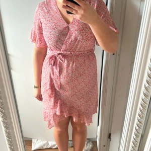 Rosa blommig klänning - Charmig rosa blommig klänning med omlott-design och knytband i midjan. Klänningen har korta ärmar med volangdetaljer och en v-ringad hals. Perfekt för somriga dagar.