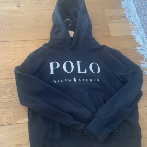 Svart hoodie från Ralph Lauren - Säljer en svart hoodie från Ralph Lauren med stor POLO-logga på framsidan. Tröjan har en klassisk design med huva och en stor ficka framtill. Perfekt för en avslappnad stil.den är i storlek XL i kids