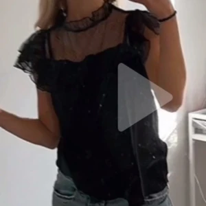 Svart blus med volangdetaljer - Snygg svart blus med volangdetaljer och transparenta partier. Blusen har korta ärmar och en hög krage med spets. Perfekt för en stilren look.