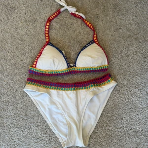 Vit bikini från Hunkemöller - Säljer en snygg vit bikini från Hunkemöller med färgglada virkade detaljer i rött, gult och blått. Bikinin har knytband i nacken och en bekväm passform. Perfekt för sommarens alla badäventyr! 🌞