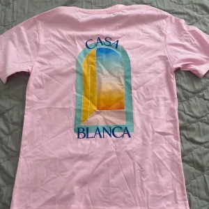 Casa Blanca rosa t-shirt med tryck - Snygg rosa t-shirt från Casa Blanca med stort färgglatt grafiskt tryck på ryggen och blå text. Klassisk passform och korta ärmar, perfekt för dig som gillar statement-plagg. Mjuk och skön bomull som känns fräsch hela dagen. Priset kan diskuteras 