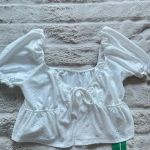 Vit blus med puffärm och knyt - Superfin vit blus med broderade detaljer, puffiga korta ärmar och söt knytning framtill. Blusen har en lätt volang nedtill och romantisk känsla, perfekt till jeans eller kjol. Luftigt bomullstyg med diskret hålmönster.
