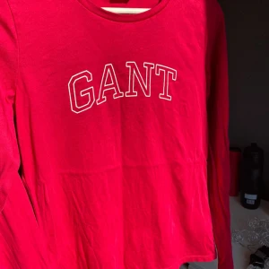 Röd långärmad tröja från GANT - Snygg röd långärmad tröja från GANT med vit broderad logga framtill. Klassisk rund halsringning och mjukt bomullsmaterial som är skönt mot huden. Perfekt för dig som gillar stilrena och sportiga plagg med tydlig logga.