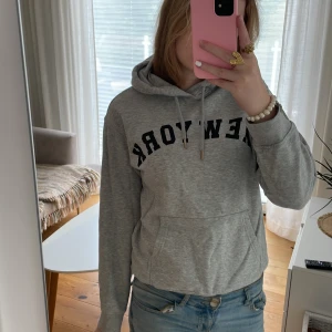 Grå hoodie med New York-tryck - Snygg grå hoodie med svart New York-tryck över bröstet. Tröjan har huva med snörning och en stor magficka framtill. Perfekt för en avslappnad och cool stil. Passar till jeans eller mjukisbyxor.
