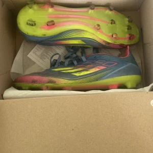 Adidas F50 fotbollsskor blå/gul - Säljer ett par Adidas F50 fotbollsskor i blått med neon-gula och rosa detaljer. Skorna har snörning och mönstrad ovandel, samt dobbar för bra grepp på planen. Perfekta för dig som vill sticka ut på fotbollsplanen.