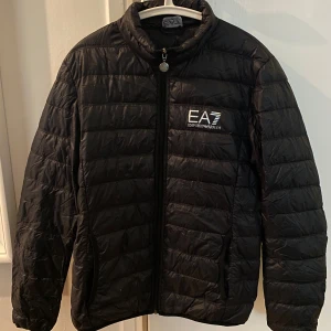 Svart dunjacka EA7 Emporio Armani - Helt ny svart dunjacka från EA7 Emporio Armani med stilren design och logga på bröstet. Jackan har quiltad look, dragkedja framtill och hög krage. Perfekt för dig som vill ha en lätt och snygg jacka med sportig vibe. NYPRIS : 2100kr. Pris går att diskutera 