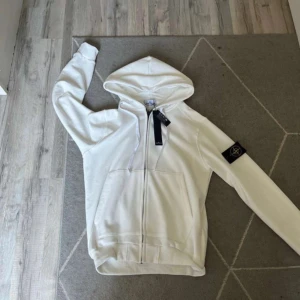 Vit hoodie från Stone Island - Vit hoodie från Stone Island med dragkedja och klassisk logga på ärmen. Tröjan har huva. Perfekt för dig som gillar stilren streetwear och vill ha något som sticker ut med ikonisk badge.