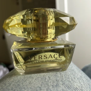 Versace Yellow Diamond parfym 50ml - Versace Yellow Diamond Eau de Toilette i en lyxig glasflaska med diamantformad gul kork. Flaskan rymmer 50 ml och har en elegant, transparent design med guldiga detaljer. Doften är fräsch och feminin, perfekt för dig som gillar exklusiva parfymer.
