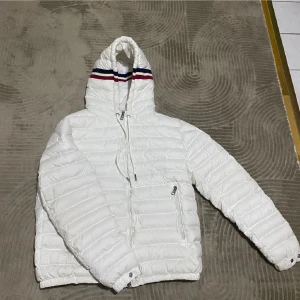 Vit pufferjacka från Moncler - Säljer en snygg vit pufferjacka från Moncler med huva och dragkedja. Jackan har ett stilrent randigt mönster i rött och blått på huvan. Perfekt för kyliga dagar.