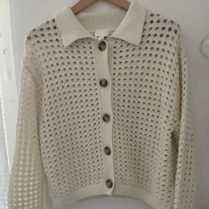 Vit stickad kofta från H&M med knappar - Säljer en vit stickad kofta från H&M i storlek S. Koftan har ett hålmönster, stora bruna knappar framtill och klassisk krage. Långärmad och perfekt för lager-på-lager. Superfin till både jeans och kjol!