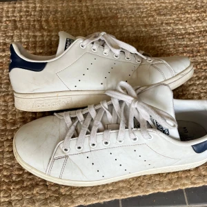 Adidas Stan Smith vita sneakers - BRA BEGAGNAT SKICK! Klassiska Adidas Stan Smith sneakers i vitt skinn med marinblå detalj på hälen och perforerade ränder på sidorna. Skorna har rund tå, platt sula och vita snören. Perfekt för dig som gillar en clean och tidlös look.