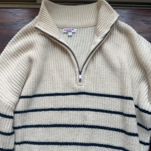 Randig stickad tröja från Jennyfer - Beige stickad tröja från Jennyfer med svarta horisontella ränder. Tröjan har hög krage, half zip-dragkedja och långa ärmar. Perfekt för dig som gillar en chill och klassisk vibe. Materialet känns rejält och mysigt. Står L men sitter bra på XS-S-M