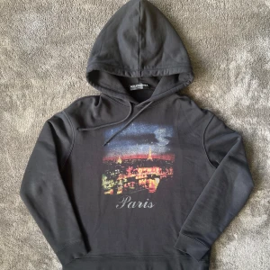 Svart Balenciaga hoodie med Paris-tryck - Svart hoodie från Balenciaga med stor huva och snörning. Framsidan har ett färgstarkt tryck av Paris skyline och texten 'Paris' i silver. Mjuk bomullskvalitet och Balenciaga-logga på ryggen. Perfekt statement-plagg för dig som gillar streetstyle. Storlek S pasar M