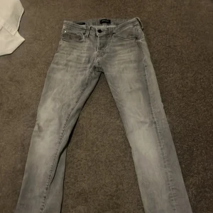 Grå slim jeans Jack & Jones 27/30 - Snygga ljusgrå skinny jeans från Jack & Jones i storlek 27/30. Jeansen har klassisk femficksdesign, dragkedjegylf och en tvättad look som ger en cool vibe. Perfekta till sneakers och hoodie för en avslappnad stil.