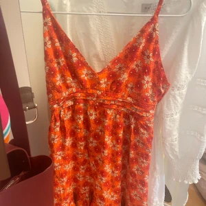 Orange blommig kort sommarklänning - Superfin orange kort klänning med vita och röda blommor, smala axelband och v-ringning. Klänningen har en lätt volang nertill och smock i ryggen för skön passform. Perfekt för varma dagar och ger en färgglad vibe. Knappt använd🩷