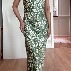 Grön blommig långklänning från Zara - Supersnygg långklänning från Zara i grönt och vitt med blommigt mönster. Klänningen har ärmlös design och figurnära passform som ger en elegant siluett. Perfekt för dig som vill sticka ut med ett unikt print och fräsch färg.