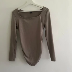 Beige långärmad topp med fyrkantig ringning - En stilren beige långärmad topp med fyrkantig ringning och lätt rynkade detaljer baktill. Toppen har en smickrande passform och är tillverkad i ett mjukt, stretchigt material som känns skönt mot huden. Perfekt att styla med jeans eller kjol för en clean look.