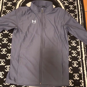 Grå träningsbyxor från Under Armour - Säljer ett par grå träningsbyxor från Under Armour i storlek XL för ungdom. Byxorna har en enkel och sportig look med resår i midjan och Under Armour-logga på låret. Perfekta för träning eller chill, tillverkade i ett mjukt och stretchigt syntetmaterial.