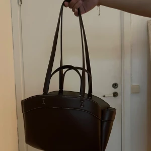 Mörkbrun axelväska i skin från Zara - Snygg mörkbrun axelväska i skinn med stilrena vita sömmar och både korta och långa handtag. Väskan har en rymlig insida med innerfack som stängs med dragkedja, perfekt för att hålla ordning på dina grejer. Enkel och klassisk design som passar till allt.