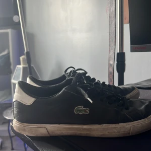 Svarta sneakers från Lacoste - Svarta sneakers från Lacoste med klassisk krokodillogga på sidan. Skorna har vita detaljer vid hälen, svarta skosnören och platt sula. Tillverkade i skinn för en stilren look som funkar till det mesta.