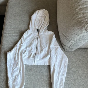 Vit croppad hoodie från H&M Divided - Säljer en vit croppad hoodie från H&M Divided i storlek XS. Tröjan har dragkedja framtill, huva och långa, breda ärmar. Perfekt för en chill och avslappnad stil. Tillverkad i mjuk bomullsmix som känns skön mot huden.