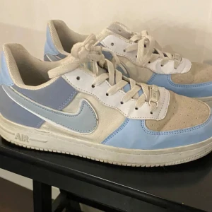 Nike Air Force 1 i ljusblå och beige - Nike Air Force 1 sneakers i en fräsch kombo av ljusblått, beige och vitt. Klassisk siluett med snörning, rund tå och platt sula. Ovandel i mocka och syntet, med ikonisk Swoosh i ljusblått på sidorna. 