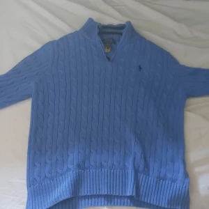 Blå kabelstickad tröja Polo Ralph Lauren - Kabelstickad blå tröja från Polo Ralph Lauren med halv dragkedja och hög krage. Klassisk design med ribbade muddar och diskret broderad logga på bröstet. Perfekt för lager-på-lager och snygg till jeans eller chinos. Går att diskutera pris. Knappt använd 5 gånger. Längd 150-167