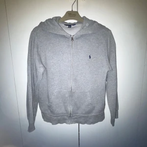 Grå zip från Polo Ralph Lauren - Klassisk grå hoodie från Polo Ralph Lauren med dragkedja framtill, huva och känguruficka. Liten blå broderad logga på bröstet. Perfekt för chill dagar och enkel att matcha med jeans eller joggers.