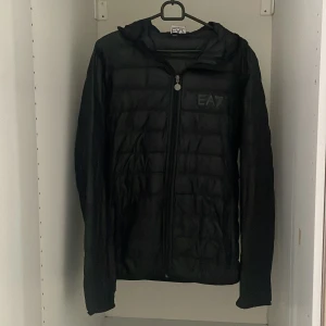 Armani/EA7 jacka - Svart pufferjacka från EA7 med diskret logga på bröstet. Jackan har huva, hel dragkedja och quiltad design. Perfekt för dig som gillar en stilren look med sportig touch. Lätt och smidig att bära, passar till många outfits.