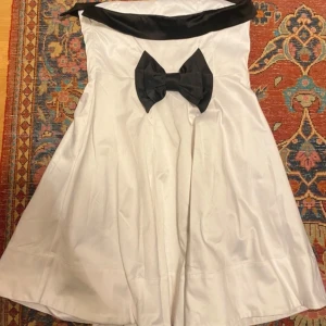 Vit axelbandslös klänning med svart rosett - Köpt på vinted men aldrig använd av mig , bilder tagna från förra säljaren. Supersöt axelbandslös vit klänning med svart kant upptill och stor svart rosett där bak. Klänningen har en utsvängd kjol och är tillverkad i ett glansigt material som ger en lyxig känsla. Den har även en dragkedja på sidan som gör den lätt att sätta på. Den har ett litet svart sträck på sig som man inte lägger märke till.