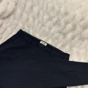 Mörkblå randiga kostymbyxor Only & Sons - Snygga mörkblå kostymbyxor från Only & Sons med tunna vita ränder. Klassisk passform med bälteshällor, knapp och dragkedja fram samt fickor bak. Perfekta för en clean och stilren look.