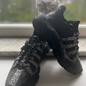 Svarta Versace Chain Reaction sneakers - Svarta Versace Chain Reaction sneakers med chunky sula, vita grafiska detaljer och snörning. Skorna har mönstrad ovandel i mesh och skinn, samt Versace-logga på sidan och bak. Snygga kontrastsömmar och unikt grekiskt mönster på sulan och tån.
