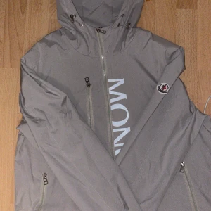 Grå vindjacka från Moncler med huva - Snygg grå vindjacka från Moncler med stor logga framtill och klassisk patch på ärmen. Jackan har huva med snörning, flera dragkedjefickor och en clean look. Perfekt för dig som gillar stilrena och exklusiva plagg med sportig vibe.