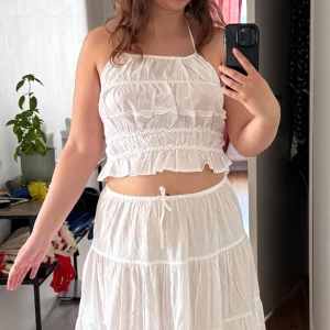 Vit volangig linnetopp från NA-KD - Supersöt vit linnetopp med halterneck från asos med volanger och smockdetaljer. Toppen har tunna knytband i nacken och är croppad med luftig känsla, perfekt för varma dagar. Kommer med en matchande maxi kjol. Storlek M, men passar även S och L då den är i stretch. Helt ny aldrig använd! Frakt tillkommer 