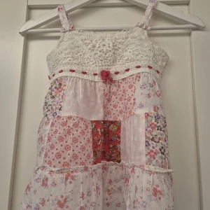 Blommig sommarklänning med spets - Supersött linne  med virkade spetsdetaljer upptill och smala axelband. Linner har patchworkmönster i rosa, rött och vitt med små blommor och en söt rosett framtill. Perfekt för varma dagar och ger en romantisk vibe.