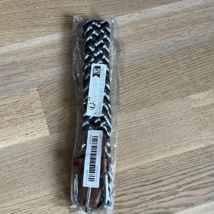 Svartvitt flätat bälte med läderdetalj - Snyggt svartvitt flätat bälte med bruna läderdetaljer vid spännet. Bältet har ett randigt mönster och passar perfekt för att ge outfiten en cool touch. Flexibelt material som gör det bekvämt att bära hela dagen.