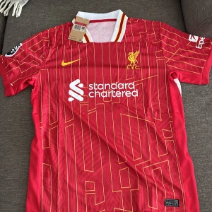 Liverpool FC röd matchtröja Nike L - Liverpool FCs officiella matchtröja från Nike i rött med gula och vita detaljer. Tröjan har korta ärmar, vit krage med gula och röda ränder, samt klubbmärke och sponsortryck på bröstet. 