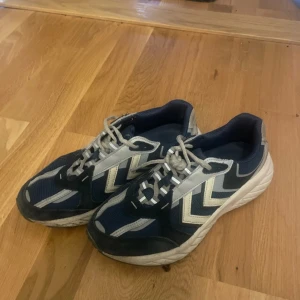 Blå och vita sneakers från Hummel - Snygga sneakers från Hummel i marinblått och vitt med klassiska pilar på sidorna. Skorna har snörning, rund tå och en bekväm platt sula. Ovandelen är i mesh och syntet. 