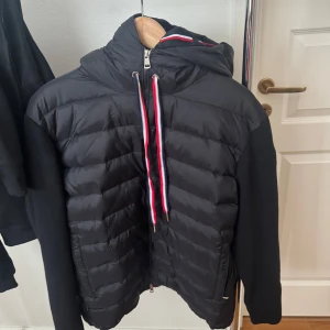 Svart cardigan från Moncler - Svart pufferjacka från Moncler med quiltad front, stickade ärmar och huva. Snygga detaljer med röd, vit och blå snörning samt dragkedja framtill. Moncler-logga på ärmen och coola metalldetaljer. Perfekt för dig som vill ha en stilren och sportig look.
