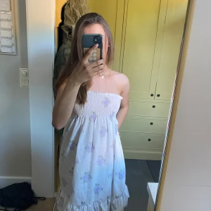 Vit blommig axelbandslös klänning - Supersöt vit klänning med lila blommor och volang nertill. Axelbandslös modell med smock upptill som sitter skönt och ger en fin passform. Perfekt för varma dagar och somriga vibes. Är i storlek xs/s och är hemma sydd💗🌸