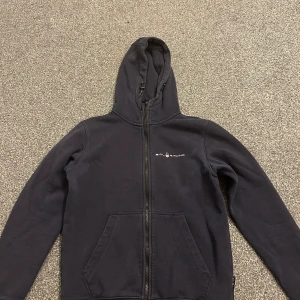 Mörkblå hoodie från Sail Racing - Hoodie från Sail Racing med dragkedja framtill och huva med snörning. Tröjan har två stora fickor på magen och diskret logga på bröstet. Perfekt för dig som gillar en clean och sportig stil.