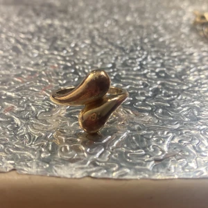 Guldig statementring med droppform - Unik ring i guldtonad metall med två droppformade detaljer som möts framtill. Ringen har en modern och organisk design som verkligen sticker ut. Perfekt för dig som gillar smycken med lite edge och cool form.