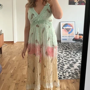 Pastellfärgad långklänning med volang - Superfin långklänning i pastellfärger. Klänningen har smala axelband, v-ringning med volangdetalj och virkade partier i midjan. Mönstrad med guldiga inslag och små blad, samt tofsdetalj framtill. Perfekt för sommarens alla tillfällen! Märket har trendat på TikTok 🛍️