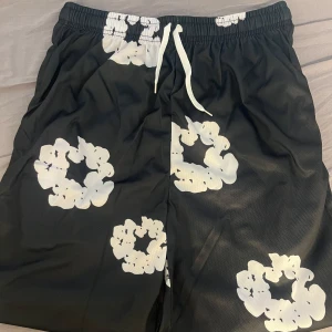 Denim tears shorts - Svarta shorts med elastisk midja och vit snörning. Shortsen har stora vita blommönster över hela tyget och är tillverkade i ett lätt, sportigt material som känns luftigt. Perfekta för en chill och trendig look under varma dagar. Shortsen är alldrig använda… tveka inte på att fråga mig vid någon fundering om shortsen…