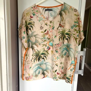 Blommig blus från House of Soul - Superfin blus med tropiskt blommönster i pastelliga färger och inslag av turkos, grönt och orange. V-ringad med korta ärmar och orange kantband. Luftig och skön, perfekt för varma dagar när du vill sticka ut lite extra.