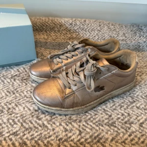 Metalliska sneakers från Lacoste - Snygga sneakers från Lacoste i metallic-brons färg med klassisk krokodillogga på sidan. Perfekta för dig som vill sticka ut med en unik färg på fötterna.