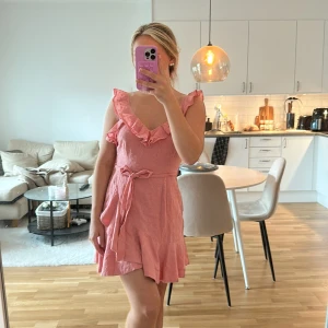 Rosa volangklänning med knytband - Supersöt rosa kortklänning med volangdetaljer längs urgingen och axlarna. Klänningen har knytband i midjan och dragkedja i sidan🌸