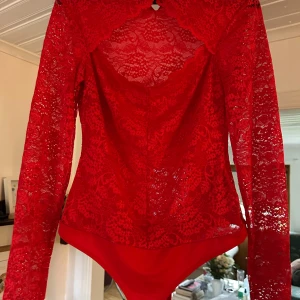 Röd spetsbody med lång ärm - Säljer en röd body i spets med långärmade ärmar och vacker blommig spetsdetalj. Toppen har knäppning i nacken med små röda knappar och en snygg öppen rygg. Perfekt för dig som vill sticka ut med en feminin och trendig look.