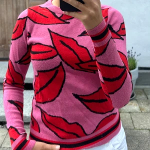 Rosa stickad tröja med rött mönster - Säljer en rosa stickad tröja med stora röda bladliknande mönster och svarta konturer. Tröjan har rund hals, långa ärmar och ribbade muddar med röd och svart rand. Perfekt för dig som gillar färg och vill sticka ut.